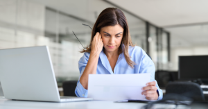 Worried serious businesswoman looking at paper bill sitting at desk in office.